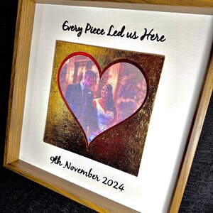 Op de afbeelding: Ingelijste trouwfoto puzzel met een hartvormige uitsparing. De puzzel toont een stel, omringd door een achtergrond van bladgoud. De tekst "Every Piece Led us Here" en "9th November 2024" staan op de lijst.
