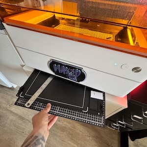 Può includere: Una stampante 3D bianca e arancione con un tappetino nero e un righello di metallo. La stampante ha il testo "MAX3D MK2" sulla parte anteriore. Il tappetino ha un motivo a griglia e segni di misurazione.