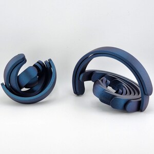 May include: Two abstract sculptures in shades of blue. One is a spherical shape with curved elements, and the other is a spiral design with a large arch. The sculptures have a metallic sheen and are set against a white background.