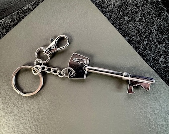 Può includere: Portachiavi a forma di chiave argentata con apribottiglie. Il portachiavi ha un design a forma di chiave con un apribottiglie all'estremità. Include una catena, un portachiavi e un fermaglio. La chiave è in metallo lucido e si trova su una superficie grigia.