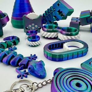 Könnte beinhalten: Eine Sammlung von 3D-gedruckten Zappelspielzeugen in schillerndem Blau, Grün und Lila. Das Sortiment umfasst eine Eidechse, Ringe, ein Zahnrad, einen Fisch und andere geometrische Formen. Ein Schlüsselanhänger ist ebenfalls enthalten.