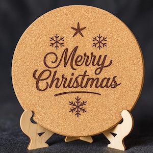 May include: A round cork coaster with the words "Merry Christmas" in a decorative font, along with snowflake and star designs. The coaster is displayed on a wooden stand, perfect for holiday decor.
