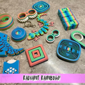 Könnte beinhalten: Eine Sammlung farbenfroher 3D-gedruckter Spielzeuge in Regenbogenfarben. Das Sortiment umfasst einen Dinosaurier, einen Gecko, ein Quadrat, eine Spirale und andere geometrische Formen. Der Text "RADIANT RAINBOW!" ist unten zu sehen.
