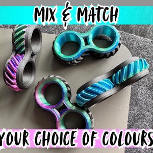 Könnte beinhalten: Mehrere 3D-gedruckte Objekte mit doppelten Löchern in verschiedenen Farben, darunter Lila, Grün und Blau, werden gezeigt. Die Objekte haben ein strukturiertes Design und sind vor einem neutralen Hintergrund platziert. Der Text oben lautet "MIX & MATCH" und unten "YOUR CHOICE OF COLOURS".