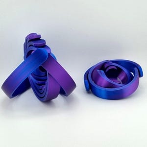 May include: Two abstract sculptures in shades of blue and purple. The sculpture on the left is a complex arrangement of curved bands, while the one on the right is a spiral design. Both are set against a white background.