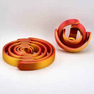 May include: Two abstract sculptures in shades of red and yellow. One is a spiral, the other a curved, open form. The colors transition from yellow to red, creating a gradient effect. The objects are set against a white background.