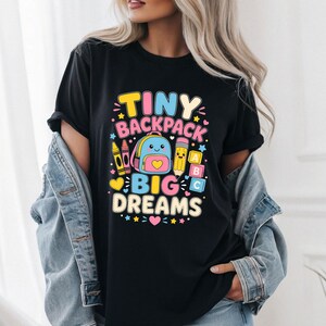 May include: A black t-shirt featuring a colorful graphic print with the text TINY BACKPACK BIG DREAMS. The design includes a cartoon backpack, a yellow crayon, a pencil, and alphabet blocks in shades of pink, yellow, blue, and white.