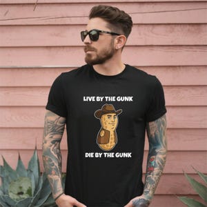 May include: Black t-shirt with the text "LIVE BY THE GUNK" above and "DIE BY THE GUNK" below a cartoon peanut wearing a cowboy hat and vest. The shirt is made of a soft material.