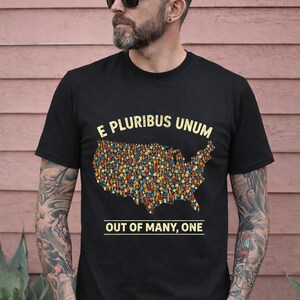 Puede incluir: Camiseta negra con la frase latina "E Pluribus Unum" y la traducción "Out of Many, One". El diseño incluye un mapa de los Estados Unidos lleno de pequeñas figuras. La camiseta la lleva una persona.