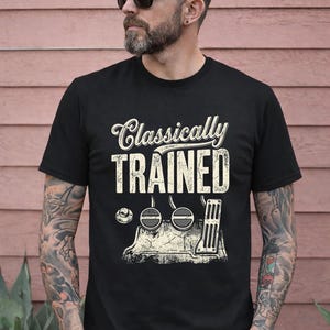 Może przedstawiać: Czarny t-shirt z napisem "Classically TRAINED" i grafiką pedałów samochodowych w stylu vintage. Osoba na zdjęciu ma na sobie okulary przeciwsłoneczne i tatuaże na ramionach.