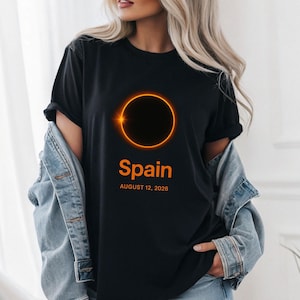 Op de afbeelding: Zwart T-shirt met een oranje ringontwerp en een donker centrum. Het woord "Spain" is in het oranje gedrukt, met daaronder "AUGUST 12, 2026". Een casual T-shirt voor dagelijks gebruik.