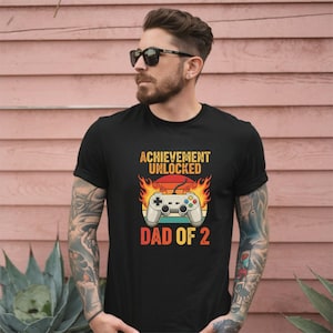 May include: Black t-shirt with the text "ACHIEVEMENT UNLOCKED" above a video game controller graphic and the text "DAD OF 2". The controller is set against a sunset and flames.