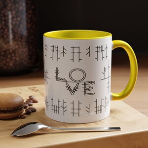 May include: White ceramic mug with a yellow interior and handle. The mug features the word "LOVE" in black, stylized lettering, surrounded by a pattern of vertical lines. A spoon and a pastry are on a wooden surface.