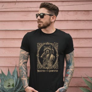 Puede incluir: Camiseta negra con un mapache tocando el violonchelo dentro de un marco dorado adornado. El texto "MAESTRO OF THE DUMPSTER" está debajo. La camiseta es de cuello redondo.