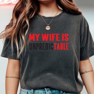 May include: Dark gray t-shirt with the text "MY WIFE IS UNPREDICTABLE" in red and black. The shirt has short sleeves and a crew neck. The model is wearing a gold necklace.