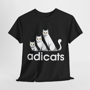 May include: Black t-shirt featuring a graphic of three stylized white cats with yellow eyes. The word "adicats" is printed in white below the cats, mimicking a well-known brand's logo. The design is centered on the shirt.