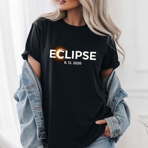 Puede incluir: Camiseta negra con la palabra "ECLIPSE" en blanco, con un círculo naranja brillante. Debajo está la fecha "8. 12. 2026" en blanco. La camiseta se lleva con una chaqueta vaquera azul.
