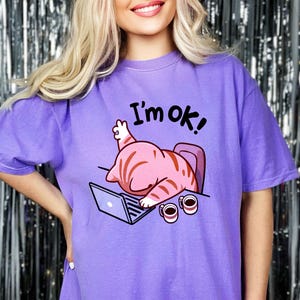 Pode incluir: Camiseta roxa com um gato de desenho animado usando um fone de ouvido sentado em uma mesa com um laptop e duas xícaras de café. O texto "I'm ok!" está impresso na camiseta.
