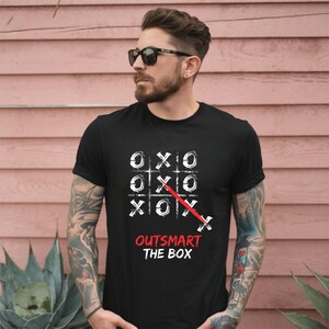 May include: Black t-shirt featuring a tic-tac-toe game design with white 'X' and 'O' symbols. A red line crosses out the winning move. The text "OUTSMART THE BOX" is printed in red below the game board.