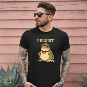Könnte beinhalten: Schwarzes T-Shirt mit dem Wort "PERSIST" in goldener Schrift über einem Cartoon-Frosch, der eine braune Mütze und eine rote Fliege trägt. Der Frosch ist grün und hellbraun.