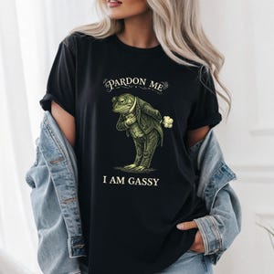 Könnte beinhalten: Schwarzes T-Shirt mit einer humorvollen Illustration eines Frosches im Anzug, mit dem Text "Pardon Me" darüber und "I Am Gassy" darunter. Der Frosch wird mit einem komischen Ausdruck dargestellt.