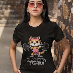Może przedstawiać: Czarny T-shirt z kreskówkowym kotem podnoszącym ciężary. Kot ma na sobie różową koszulkę bez rękawów i opaskę na głowę. Pod kotem nadrukowany jest tekst "Work hard, stay cute".
