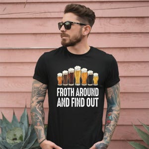 Può includere: T-shirt nero con grafica a tema birra. Il design presenta vari bicchieri di birra con schiuma e la scritta bianca "Froth Around and Find Out". La maglietta è girocollo.