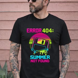 Puede incluir: Camiseta negra con un diseño retro. El texto dice "ERROR 404: SUMMER NOT FOUND". El gráfico incluye una puesta de sol pixelada y un disquete con efecto de goteo, en colores neón.