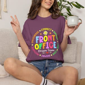 Op de afbeelding: Een bordeauxrood T-shirt met een kleurrijke afbeelding met de tekst "FRONT OFFICE Dream Team" in een cirkelvormig ontwerp met woorden als "Encourage" en "Inspire". Het shirt wordt gedragen met een spijkerbroekshort.