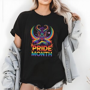 Könnte beinhalten: Schwarzes T-Shirt mit einer farbenfrohen Grafik einer gehörnten Figur mit Regenbogen-Heiligenschein und dem Text "PRIDE MONTH". Das Design verwendet leuchtende Farben und ein himmlisches Thema. Es ist ein Freizeitkleidungsstück.