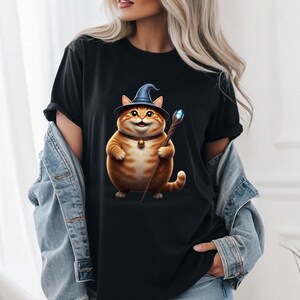 May include: Black t-shirt featuring a cartoon orange tabby cat wearing a blue wizard hat and holding a staff with a glowing blue crystal. The cat has a gold bell collar. The shirt is a casual style.