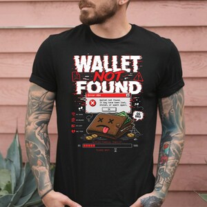 Könnte beinhalten: Ein schwarzes Kurzarm-T-Shirt mit einem Grafikdruck, der den Text "WALLET NOT FOUND" und ein "Error 404"-Fenster zeigt. Das Design enthält eine braune Geldbörse mit traurigem Gesicht, grünes Papiergeld, Goldmünzen und einen Ladebalken am unteren Rand.