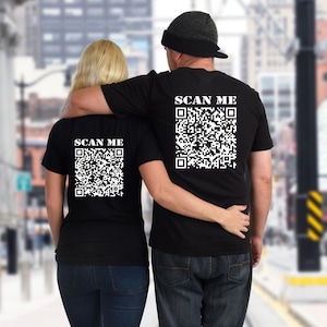 May include: Two people wearing black t-shirts with white QR codes on the back. The shirts say "SCAN ME" above the QR codes.