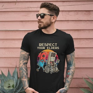Peut inclure: T-shirt noir avec le texte "RESPECT YOUR ELDERS" au-dessus d'un dessin d'une manette de jeu poussant une console portable rétro en fauteuil roulant. Le dessin a un fond de coucher de soleil.