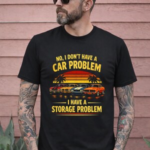 Puede incluir: Camiseta negra con un gráfico de coches clásicos y el texto "NO, I DON'T HAVE A CAR PROBLEM - I HAVE A STORAGE PROBLEM." El diseño incluye una puesta de sol y palmeras.