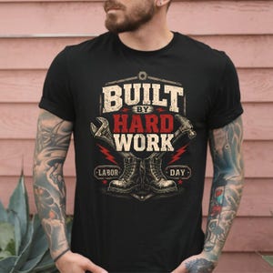 Peut inclure: Un t-shirt noir à manches courtes avec une impression graphique portant le texte BUILT BY HARD WORK et LABOR DAY. Le motif comprend des illustrations d'une clé, d'un marteau et d'une paire de bottes de travail en blanc, rouge et gris.