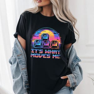 Puede incluir: Camiseta negra con un gráfico colorido que presenta las letras "W", "A", "S" y "D" en un diseño de teclado retro, sobre un fondo de puesta de sol. El texto "IT'S WHAT MOVES ME" está debajo del gráfico.