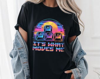 May include: Black t-shirt with a colorful graphic featuring the letters "W", "A", "S", and "D" in a retro keyboard design, set against a sunset-style background. The text "IT'S WHAT MOVES ME" is below the graphic.