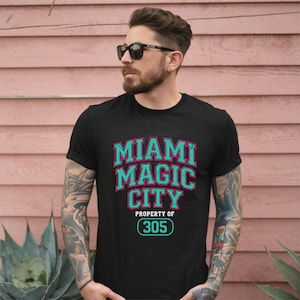 Puede incluir: Camiseta negra con el texto "MIAMI MAGIC CITY" en letras turquesas y rosas. Debajo, "PROPERTY OF 305" en blanco, con el número 305 en un círculo turquesa. La camiseta la lleva una persona.