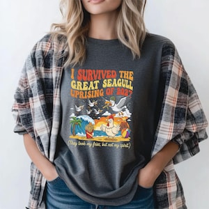 Puede incluir: Camiseta gris oscuro con un gráfico colorido que dice "I SURVIVED THE GREAT SEAGULL UPRISING OF 2027". El gráfico representa gaviotas, un pollo, papas fritas y una escena de playa. También incluye el texto "(They took my fries, but not my spirit.)".