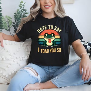 May include: Black t-shirt featuring a green frog graphic with the text "Hate to Say I Toad You So." The frog has its tongue out, set against a retro sunset design. A casual wear item.