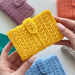 May include: A bright yellow rectangular crochet wallet featuring a textured star stitch pattern and a small central flap closure. The wallet is constructed from yarn with a consistent stitch density and a secure tab to keep the item closed.