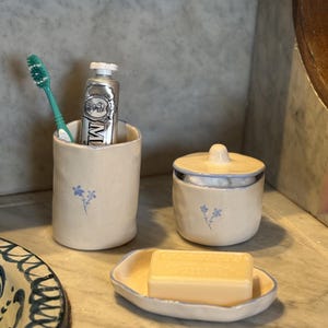 May include: A ceramic bathroom set in cream with blue floral accents, including a cylindrical toothbrush holder containing a green toothbrush and toothpaste tube, a lidded jar, and an oval soap dish holding a rectangular bar of soap.