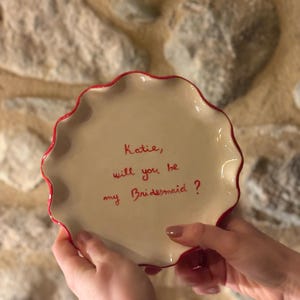 Pode incluir: Um prato pequeno de cerâmica cor de creme com borda ondulada e um fino acabamento vermelho. O centro apresenta um texto escrito à mão em vermelho que diz: Katie, will you be my Bridesmaid?