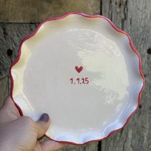 May include: A round ceramic plate with a scalloped edge and a glossy white finish. The center of the plate features a small red heart icon positioned above the date 1.1.25, which is written in a simple red font. The rim is painted in red.