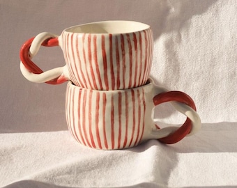 Puede incluir: Dos tazas de cerámica con rayas rojas y blancas y asas con forma de bastón de caramelo. Las tazas están apiladas una encima de la otra.