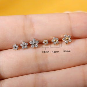 May include: Six gold-tone flower-shaped stud earrings with clear stones arranged in a row. The three largest earrings are labeled with sizes 3.5mm, 4.5mm, and 5.5mm. Each earring features a central circular element surrounded by six small clear stones.