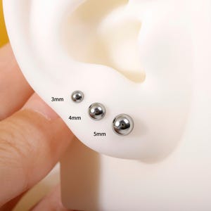 May include: Close-up of three silver ball stud earrings, 3mm, 4mm, and 5mm in size, displayed on a white ear model. The earrings are shiny and reflective, showcasing a simple, minimalist design. The sizes are labeled on the ear model.