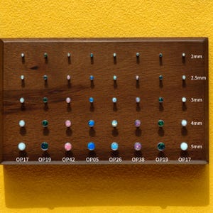 Pode incluir: Uma placa de madeira com vários brincos pequenos e coloridos. Os brincos estão dispostos em fileiras e colunas, com tamanhos que variam de 2 mm a 5 mm. Cada brinco tem uma pedra preciosa de cor diferente, incluindo branco, verde, rosa e azul.