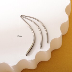 May include: Three silver curved piercing needles, each measuring 27mm in length. The needles are arranged in a curved shape on a white surface.